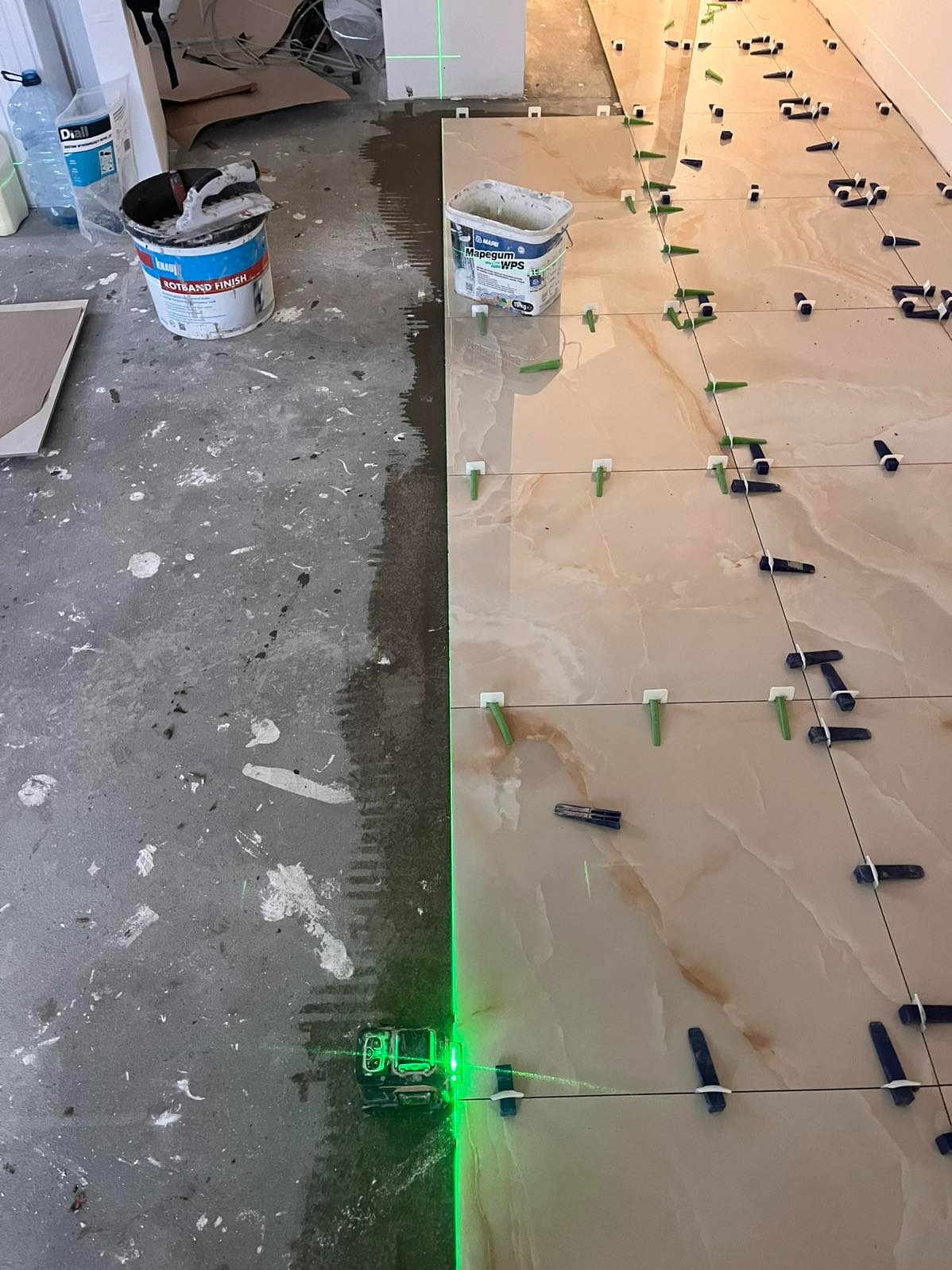 Układanie beżowych płytek z wzorem marmuru, z użyciem poziomicy laserowej i klipsów, na podłodze z widocznymi śladami zaprawy. Częściowo ułożona podłoga.