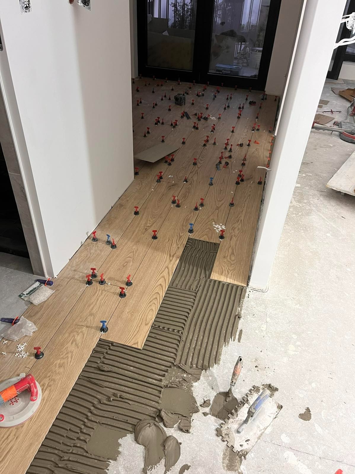 Układanie drewnopodobnych płytek podłogowych z użyciem systemu poziomowania. Klej na podłodze, narzędzia glazurnicze. Widok z góry na fragment podłogi w trakcie prac.