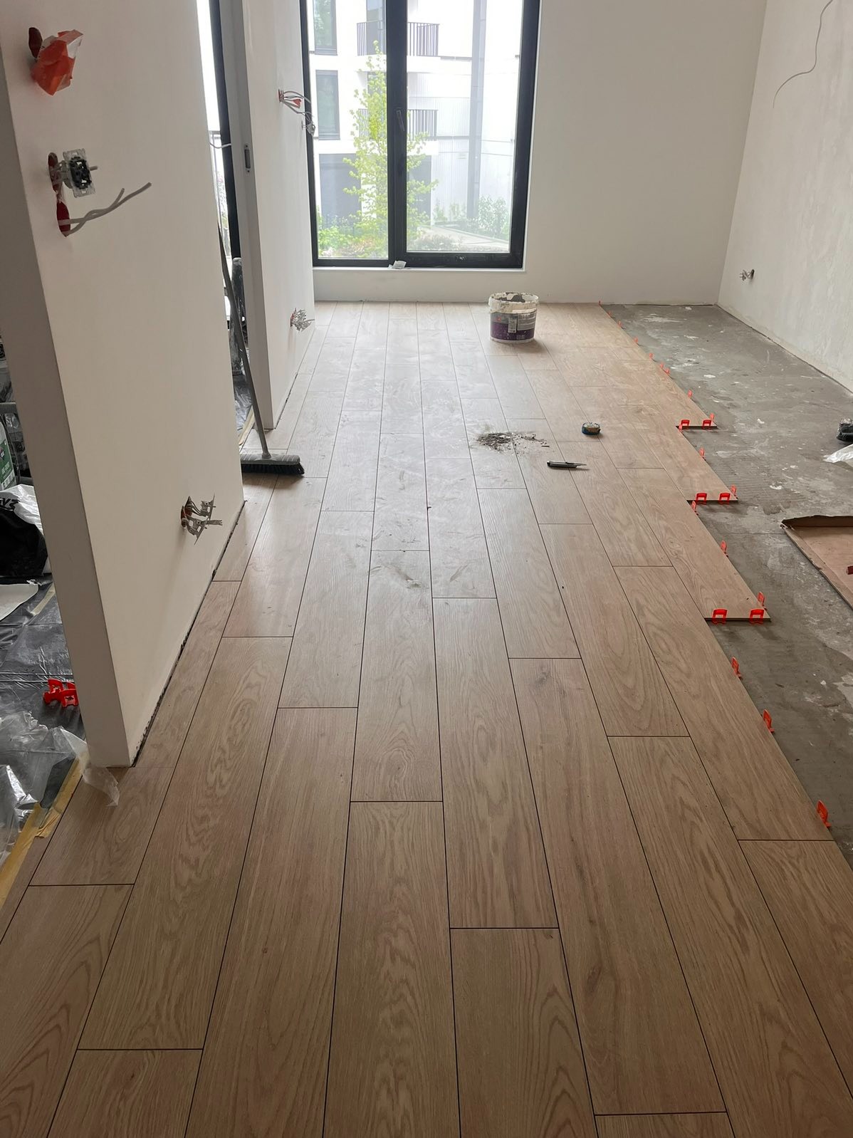 Układanie podłogi z drewnopodobnych paneli w jasnym wnętrzu. Widoczne krzyżyki dystansowe, wiadro z klejem i fragment surowej podłogi. Częściowo położona podłoga.