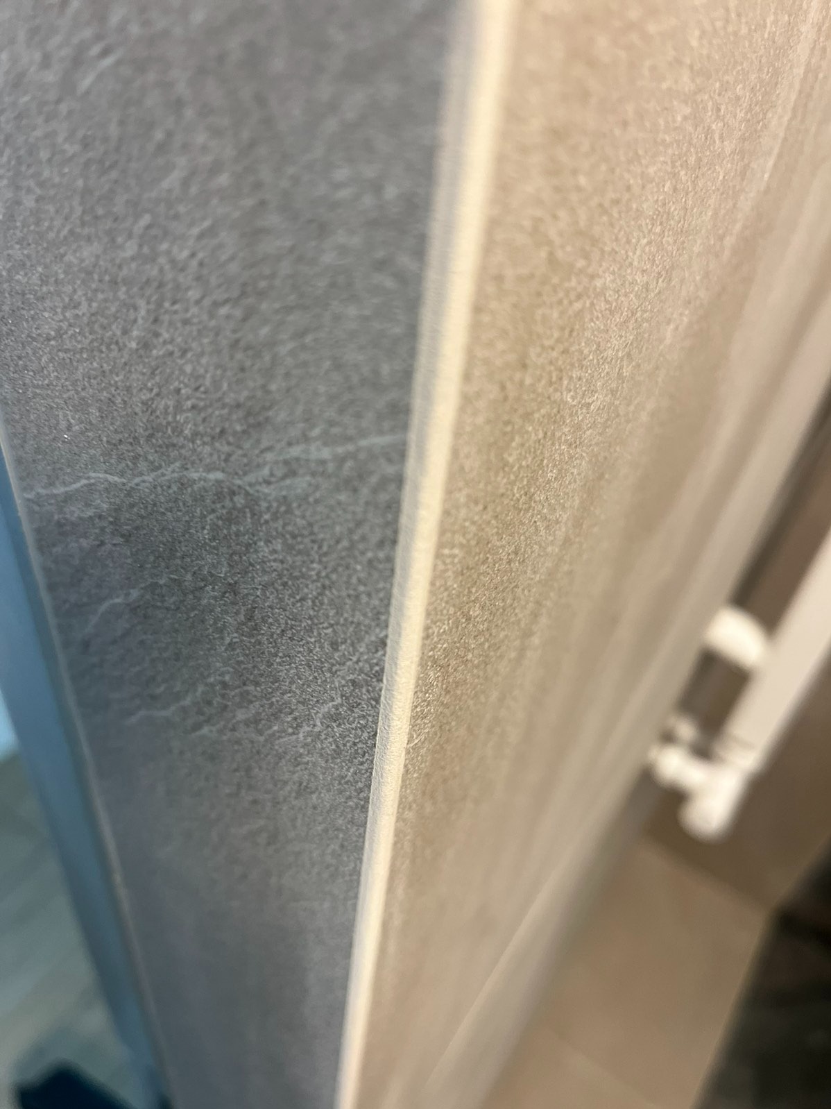 Zbliżenie na styk szarej i beżowej płytki w łazience, z widoczną fugą i fragmentem grzejnika w tle. Układ pionowy płytek. Stonowana kolorystyka.