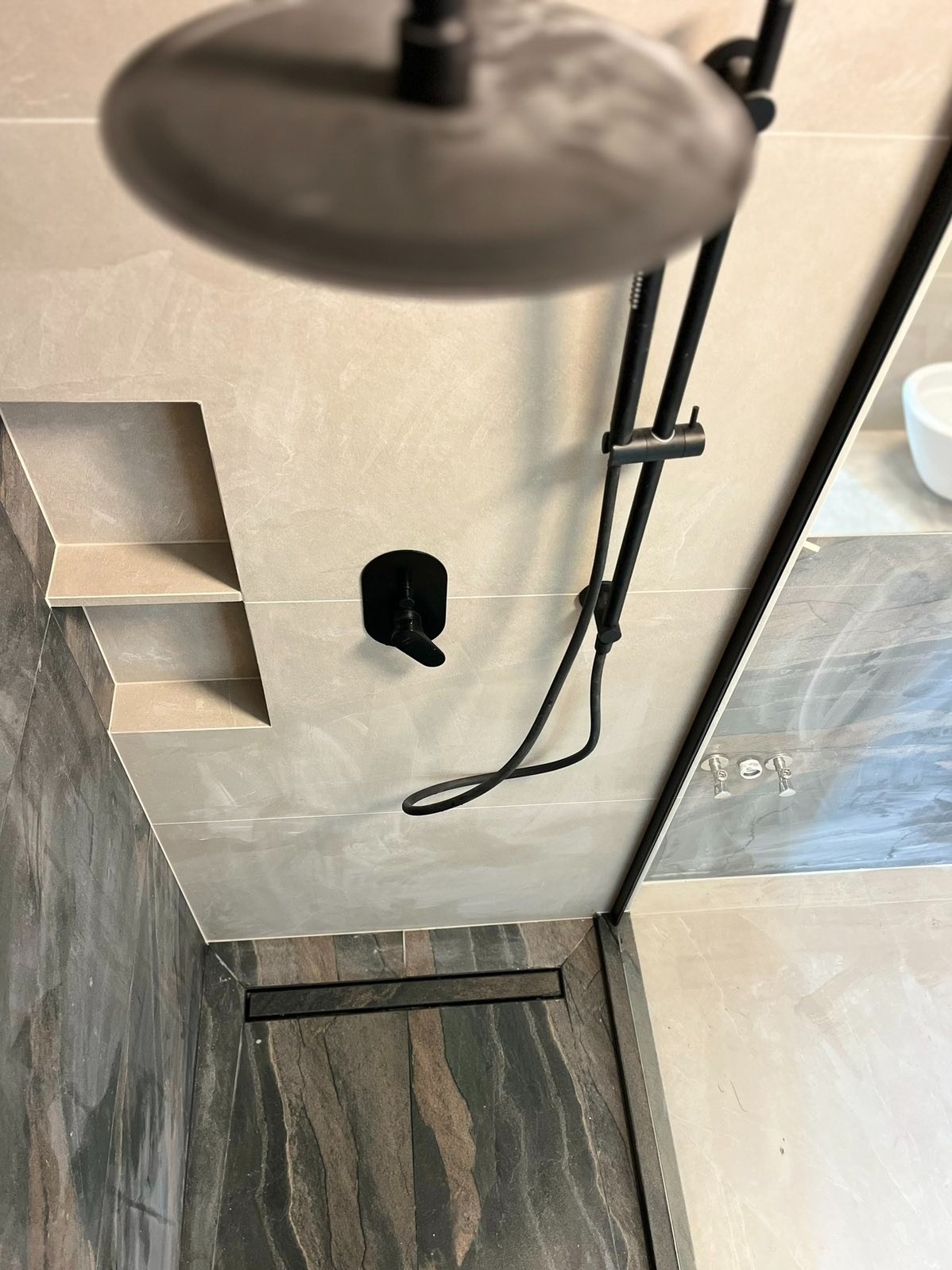 Stylowa łazienka po remoncie: deszczownica w kolorze czarnym, półki wnękowe w jasnych płytkach, odpływ liniowy i szklana kabina prysznicowa.