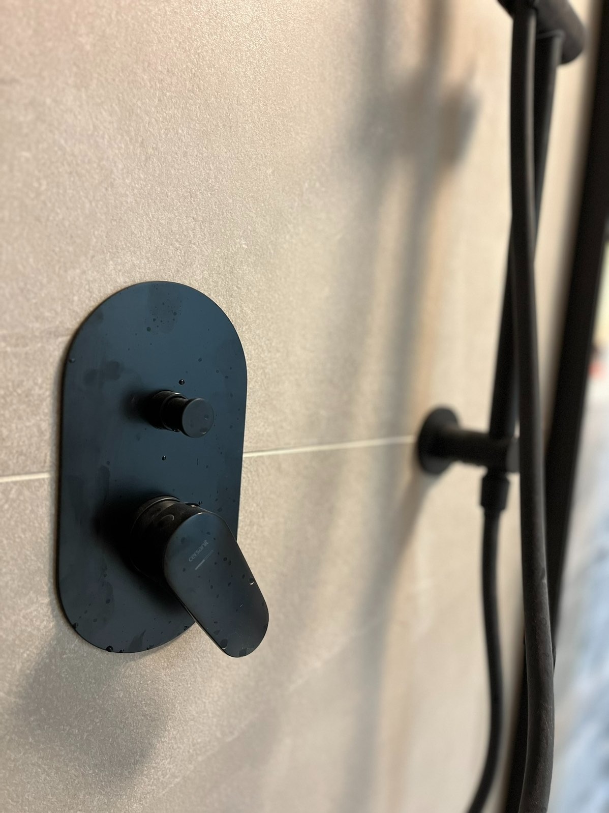 Nowoczesna, czarna bateria prysznicowa Cersanit na beżowej płytce w łazience. Minimalistyczny design, detale wykończenia, widoczne krople wody.