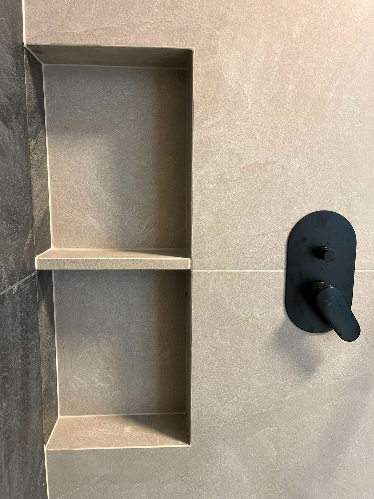 Wnęka w szarej, wykafelkowanej ścianie łazienki z czarną, nowoczesną baterią umywalkową. Minimalistyczny design i wykończenie.