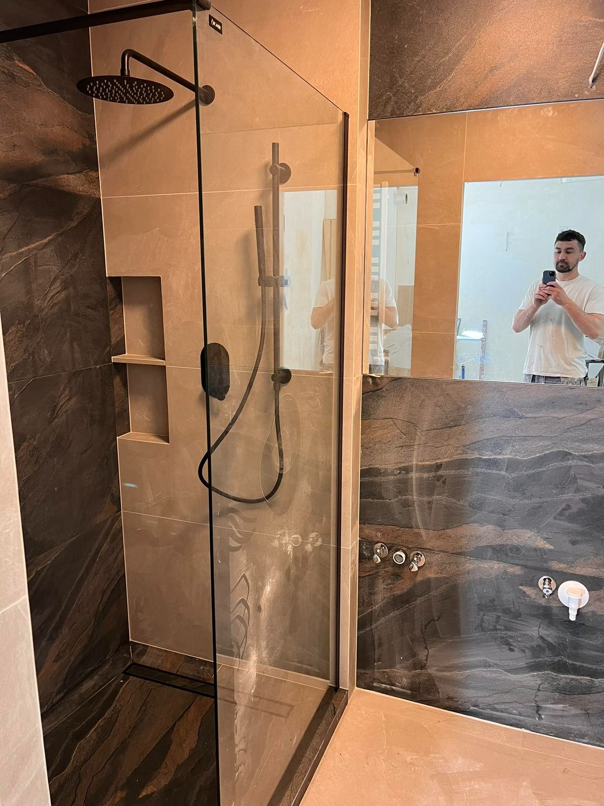 Wykończona kabina prysznicowa z deszczownicą i półkami wnękowymi, widoczna w lustrze osoba robiąca zdjęcie. Użyto płytek imitujących kamień.