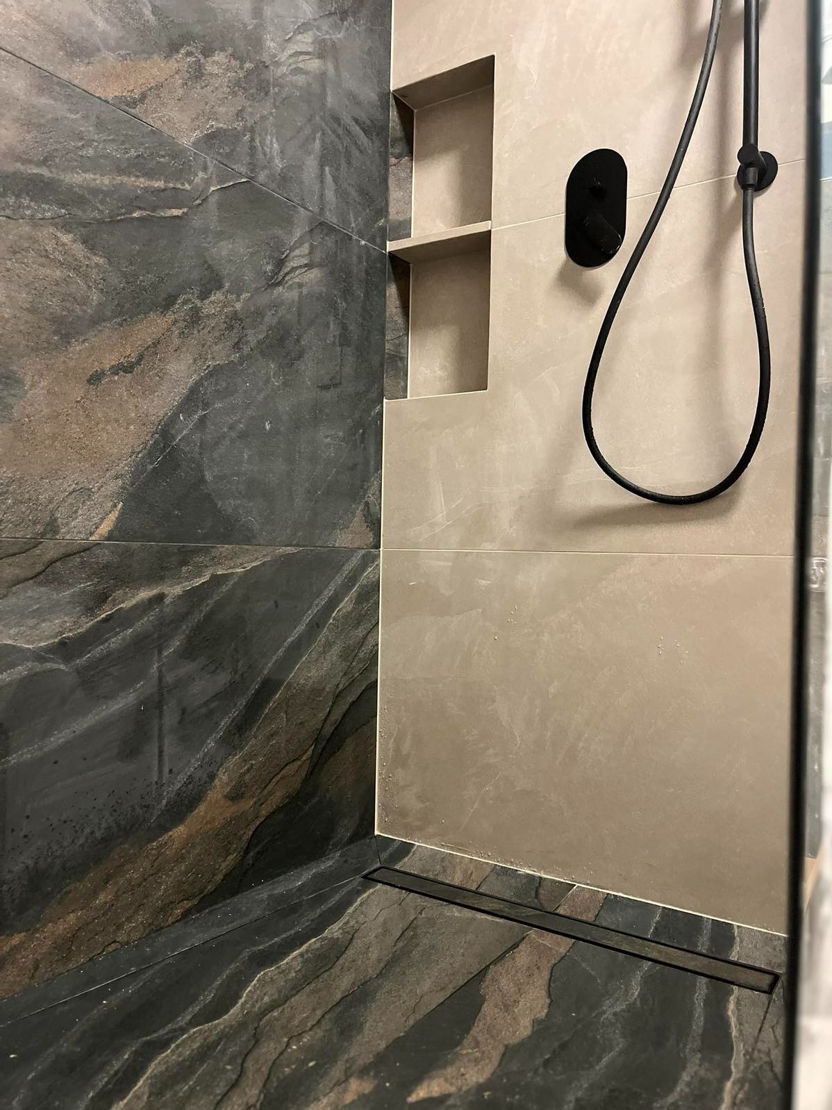 Nowoczesna kabina prysznicowa z odpływem liniowym i półkami wnękowymi. Ciemna, kamienna glazura podłogi i ściany kontrastuje z jasną ścianą z czarną armaturą.