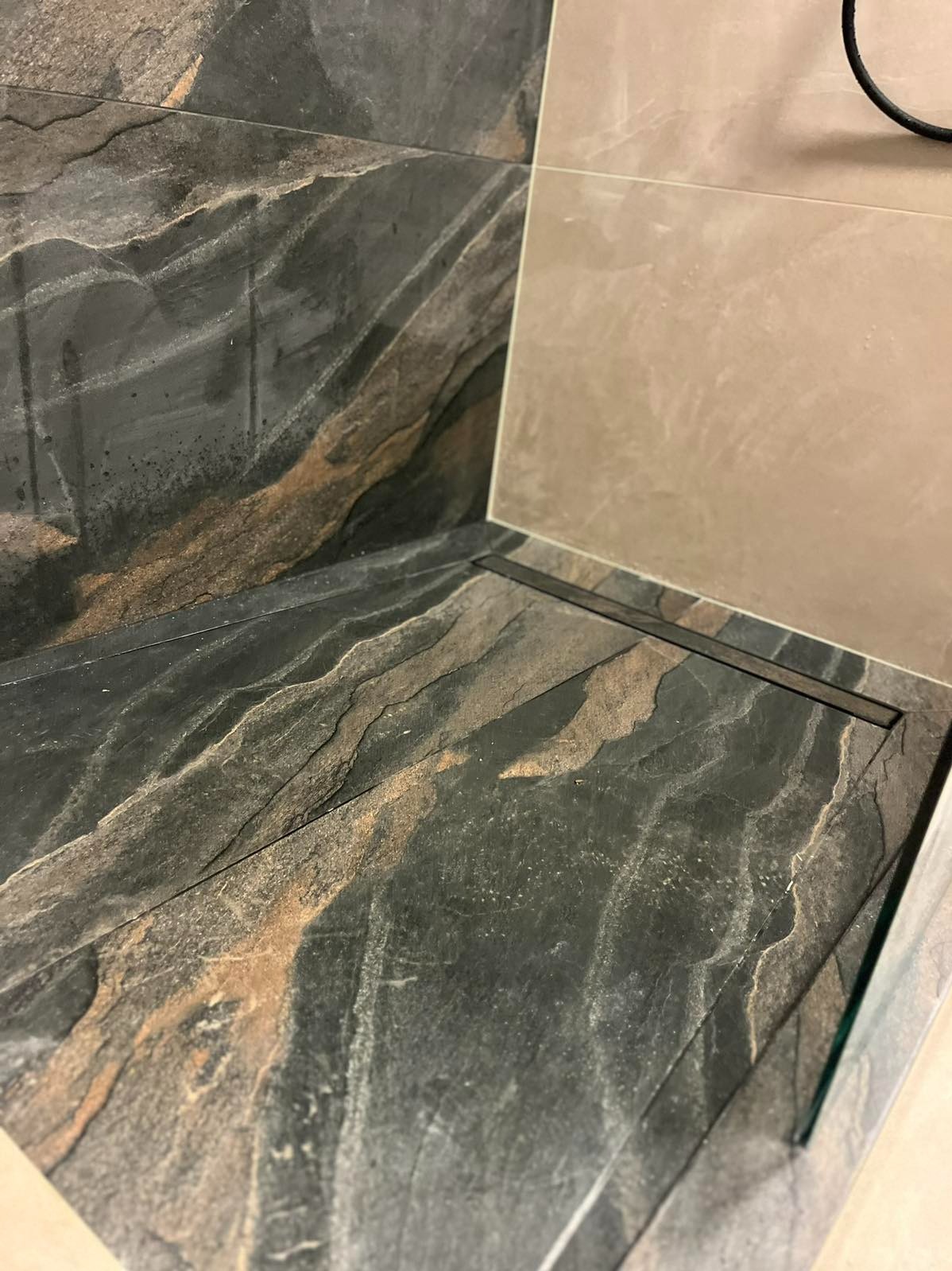 Nowoczesny odpływ liniowy w kabinie prysznicowej z czarnych płytek imitujących kamień. Jasne płytki na ścianie. Minimalistyczny design.