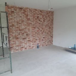 MarcusKowal1989 - Ściana wyłożona cegłą dekoracyjną w surowym wnętrzu. Widoczne rusztowanie i butelki z wodą. Oświetlenie sufitowe w trakcie instalacji.