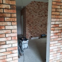 MarcusKowal1989 - Ściana wykończona czerwoną cegłą dekoracyjną, widoczna przez futrynę drzwi. Na podłodze narzędzia, w tle dalsza część ściany i sprzęt budowlany.