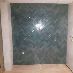 MarcusKowal1989 - Ściana wyłożona turkusową glazurą w jodełkę, z widoczną instalacją elektryczną na suficie i fragmentem drewnopodobnej podłogi, w trakcie wykańczania łazienki.