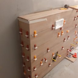 MarcusKowal1989 - Montaż płytek imitujących drewno na ścianie z użyciem klipsów poziomujących. Widoczny otwór na instalację i narzędzia glazurnicze.