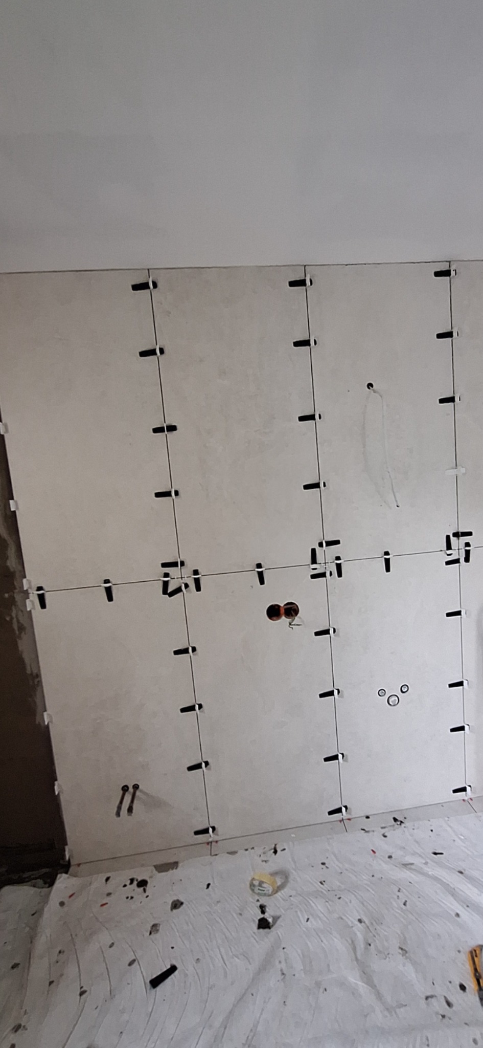 Ściana w trakcie układania płytek z krzyżykami dystansowymi. Widoczne otwory na instalacje. Podłoga zabezpieczona folią malarską.