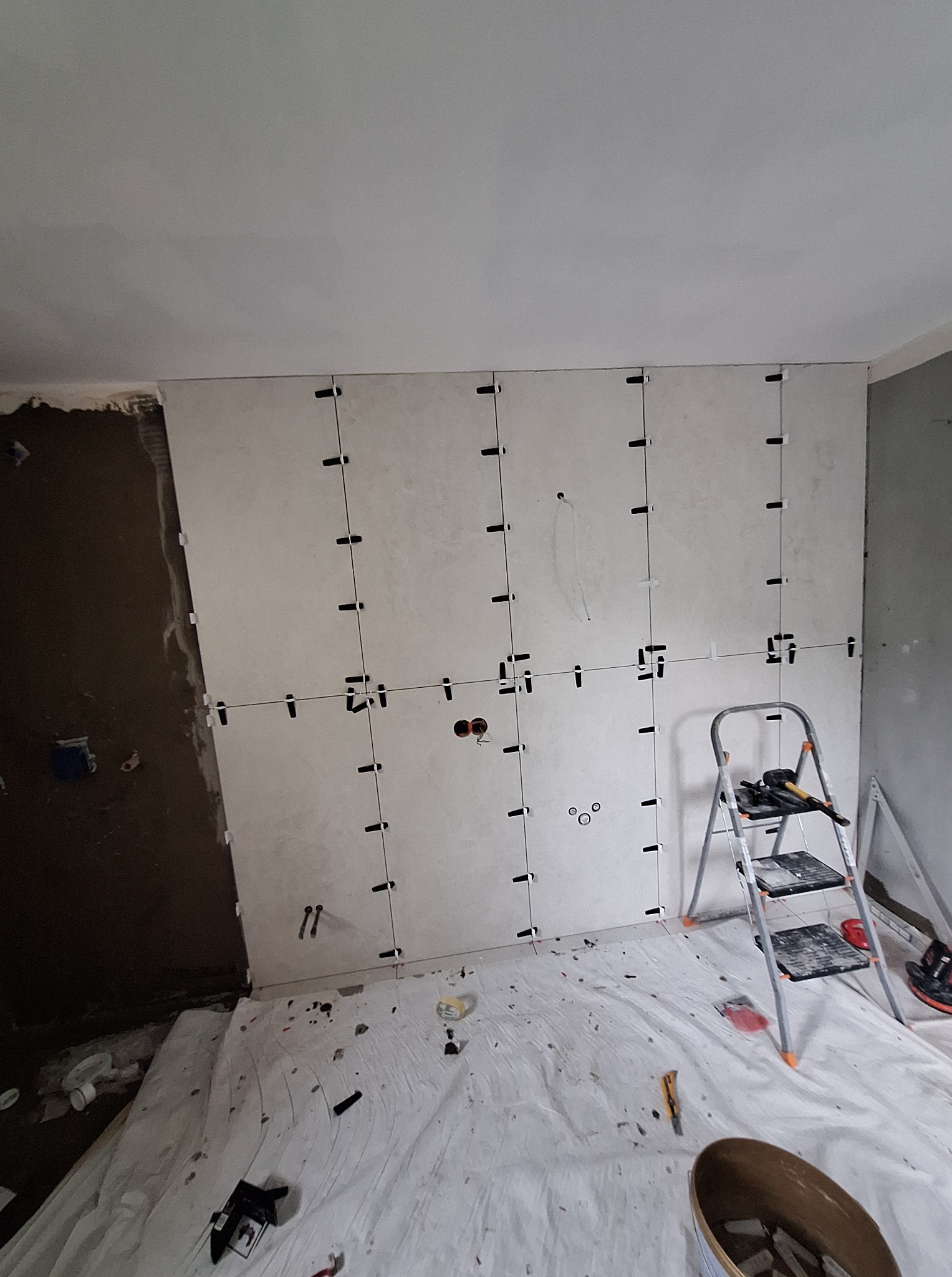 Ściana w trakcie układania płytek, widoczne krzyżyki dystansowe, drabina i narzędzia. Częściowo położone płytki w jasnym odcieniu.