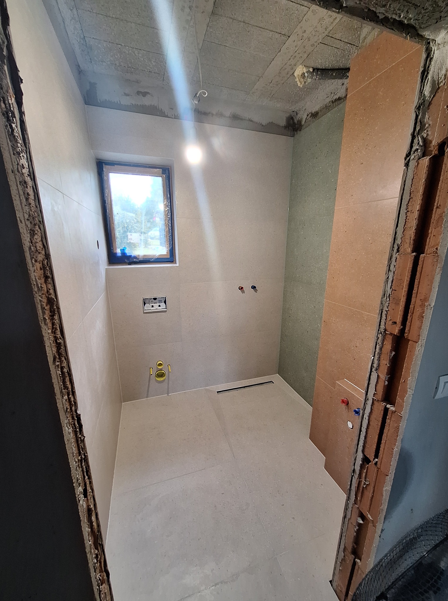 Jasna łazienka w trakcie remontu: beżowe płytki, okno z widokiem na zieleń, widoczne instalacje. Czerwona cegła przy framudze.