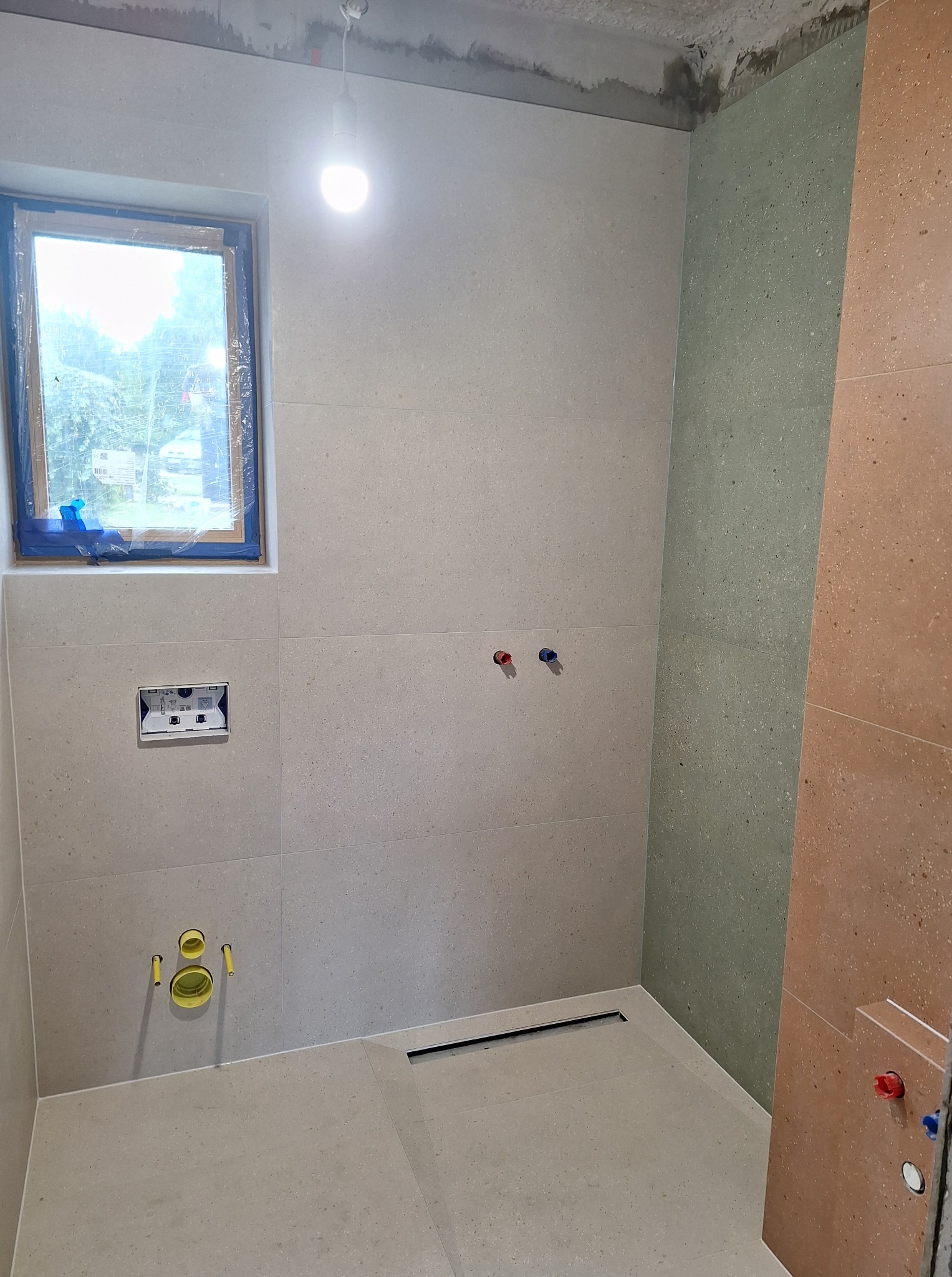 Ściany łazienki wyłożone jasnymi płytkami, widoczne przyłącza hydrauliczne i okno z folią ochronną. Ściek liniowy w podłodze. Częściowo wykończona ściana w kolorze zielonym i brązowym.