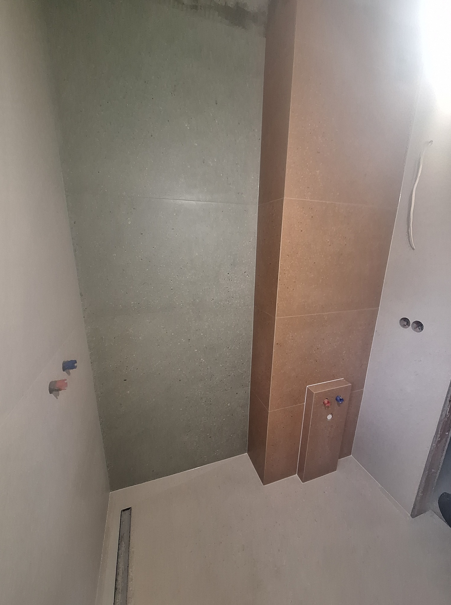 Wnętrze łazienki w trakcie prac. Widoczne szare płytki na ścianie, pionowy element obudowany brązowymi płytkami, przygotowane instalacje wodne.