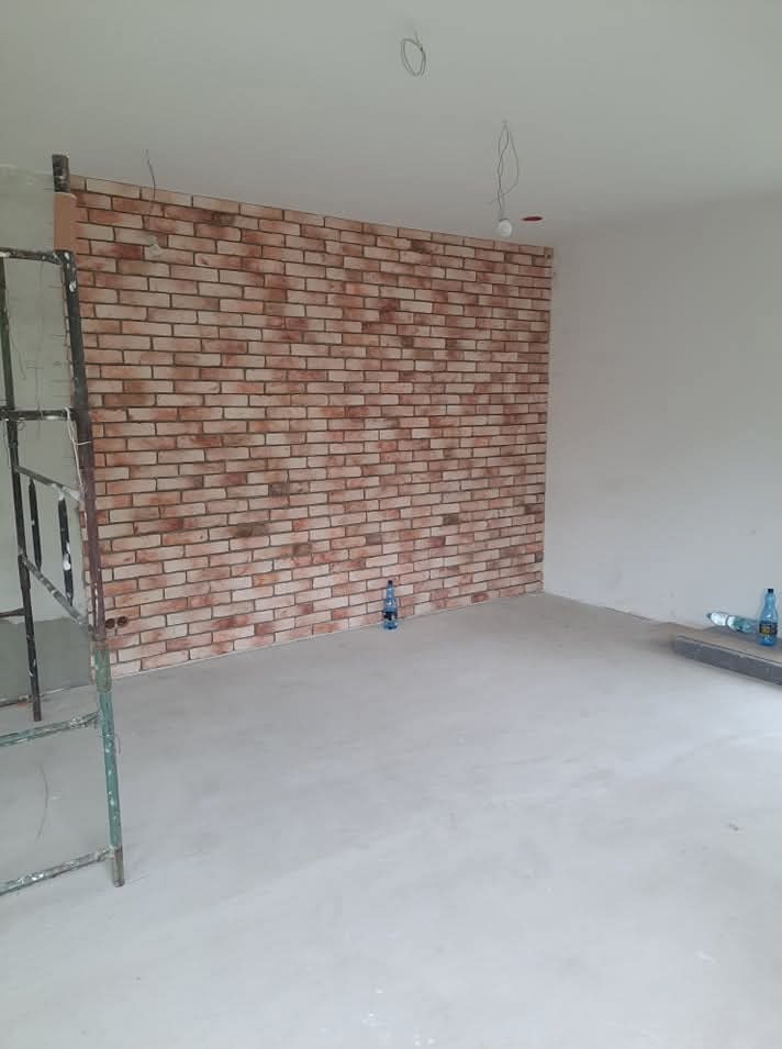 Ściana wyłożona cegłą dekoracyjną w surowym wnętrzu. Widoczne rusztowanie i butelki z wodą. Oświetlenie sufitowe w trakcie instalacji.