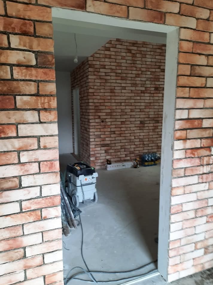 Ściana wykończona czerwoną cegłą dekoracyjną, widoczna przez futrynę drzwi. Na podłodze narzędzia, w tle dalsza część ściany i sprzęt budowlany.