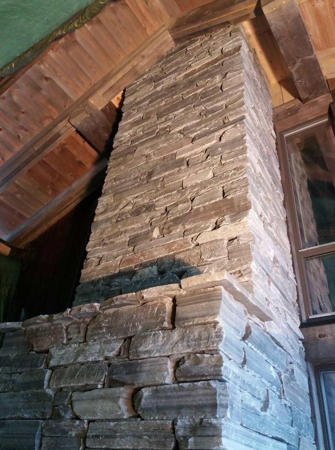 Kamienna obudowa kominka w rustykalnym wnętrzu z drewnianym stropem i oknem. Widoczna faktura kamienia i detale wykończenia.