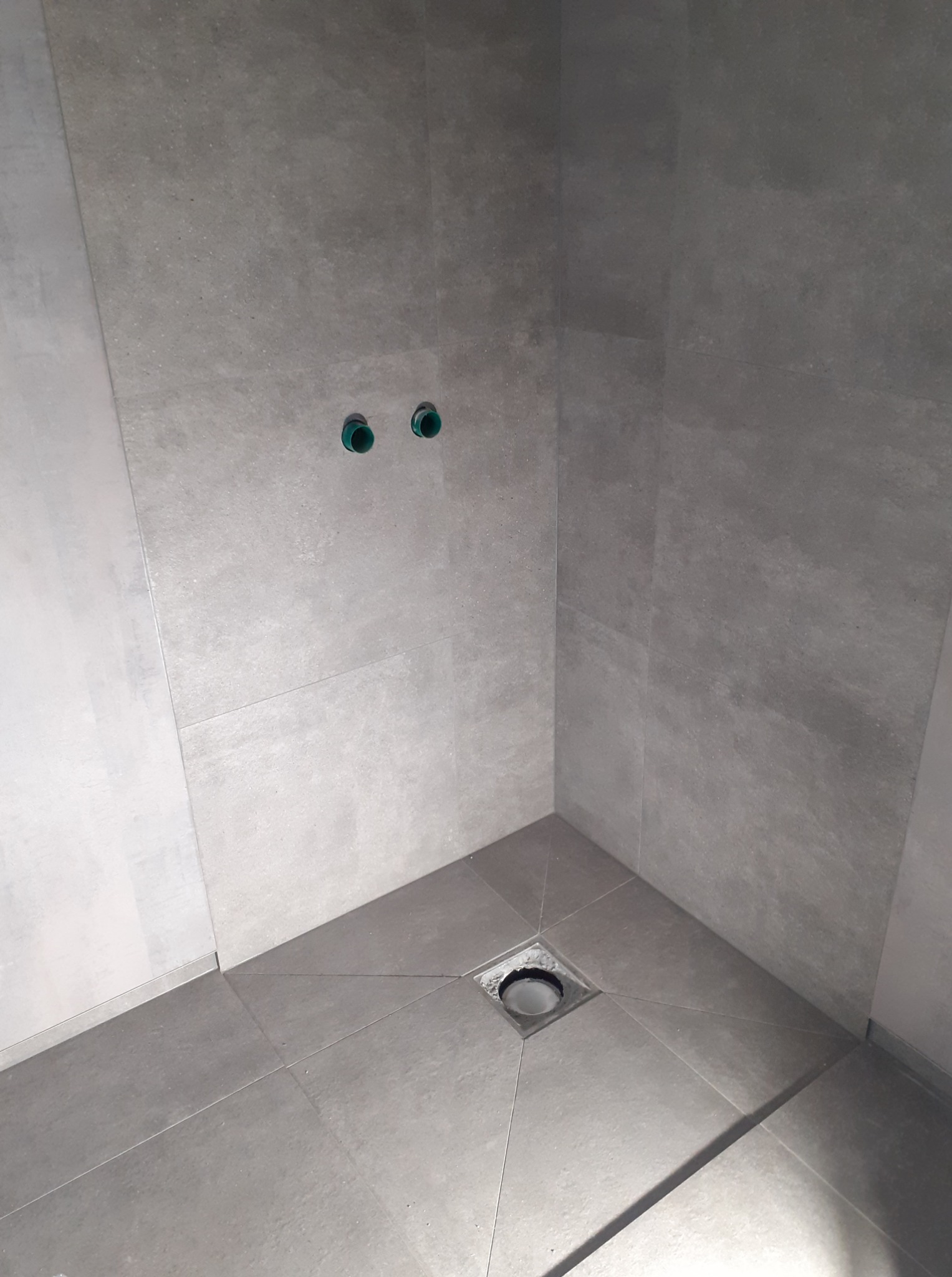 Wykończona szara łazienka z odpływem liniowym i widocznymi przyłączami wodnymi w ścianie, prezentująca minimalistyczny design i nowoczesne rozwiązanie odprowadzania wody.