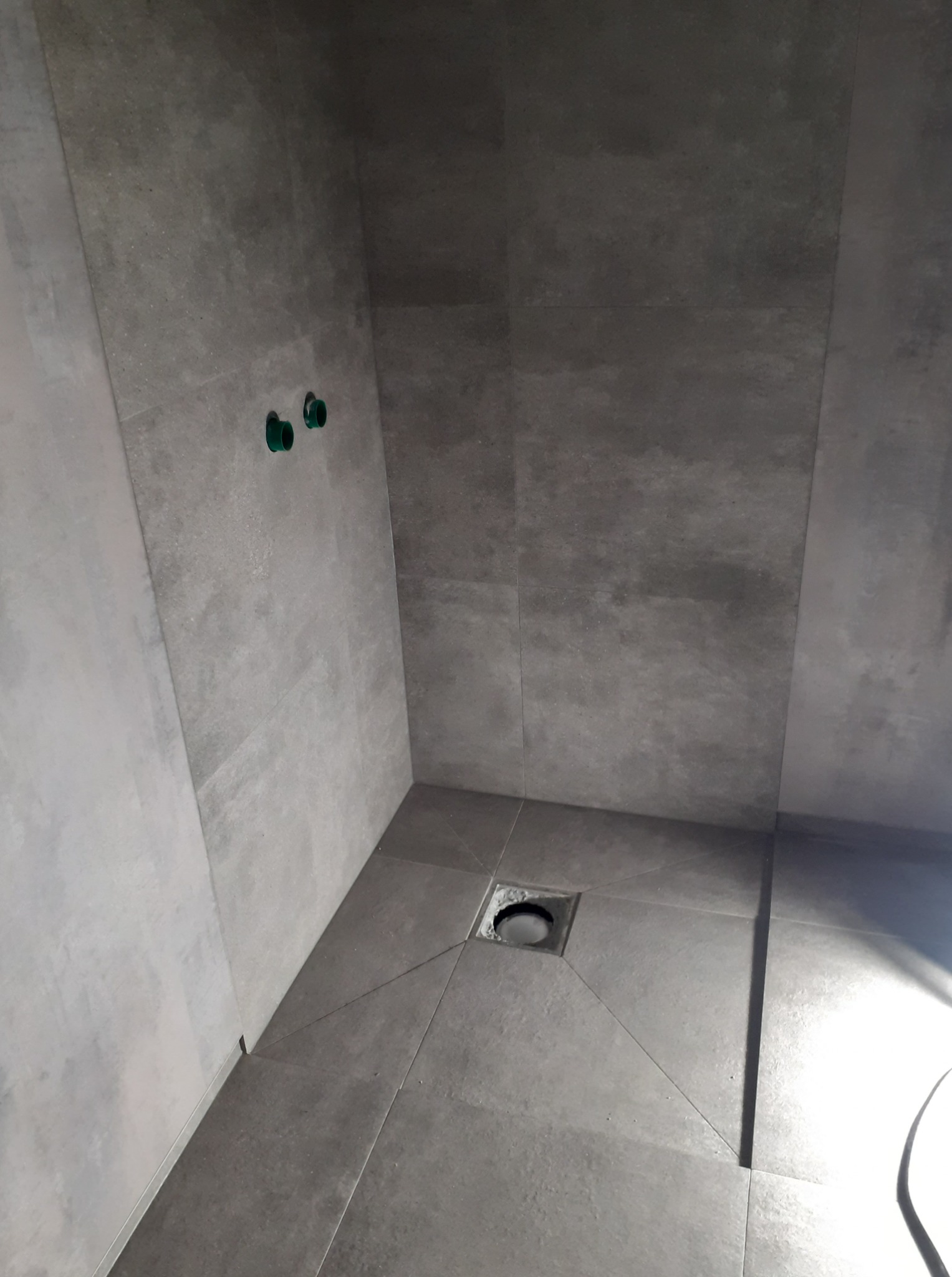 Wykończona kabina prysznicowa z szarymi płytkami wielkoformatowymi i odpływem liniowym. Widoczne przyłącza wodne. Minimalistyczny design.