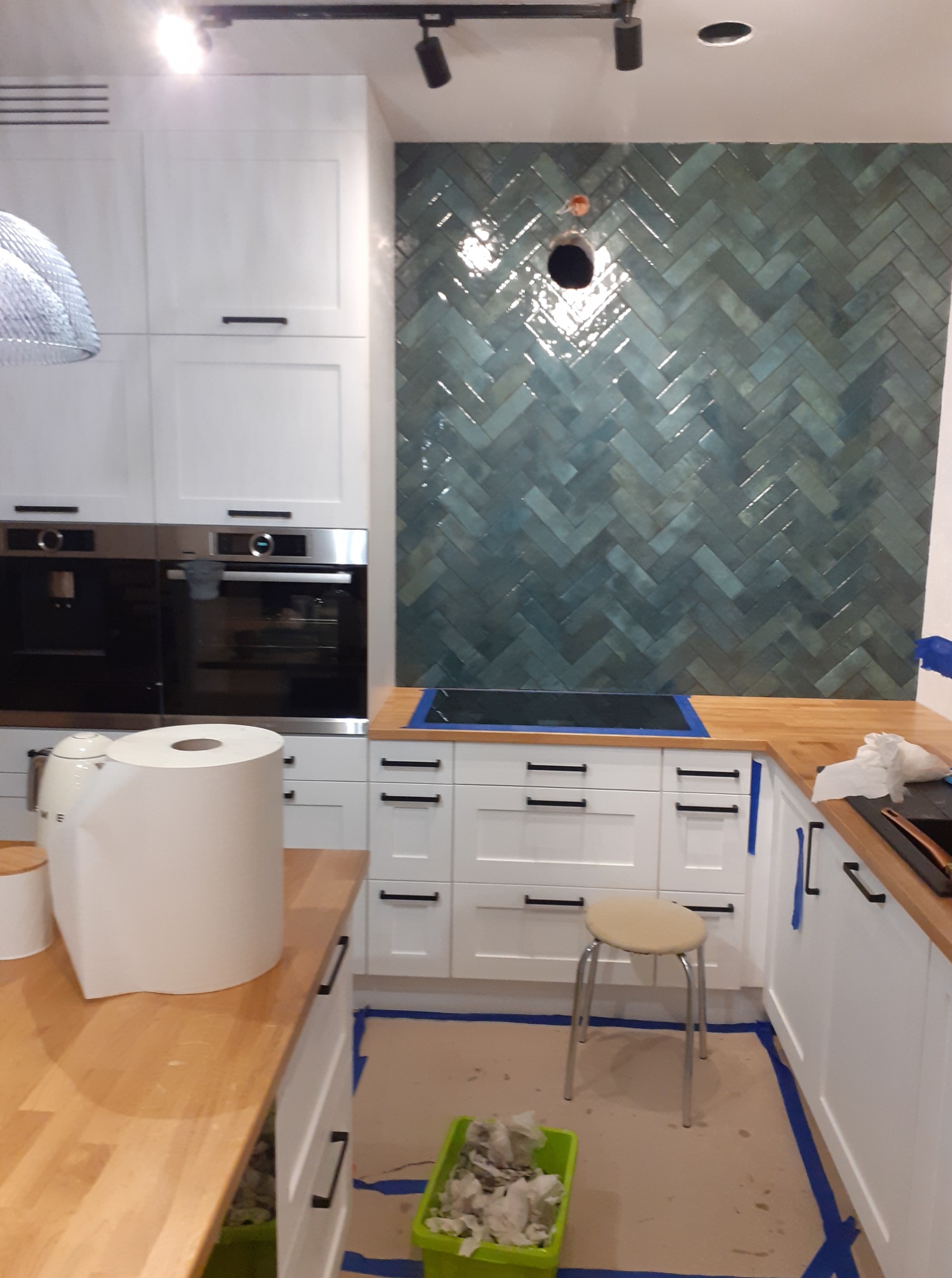 Kuchnia w trakcie wykańczania: białe szafki, drewniany blat i ściana z zielono-niebieskich płytek ułożonych w jodełkę. Widoczny otwór na gniazdko.