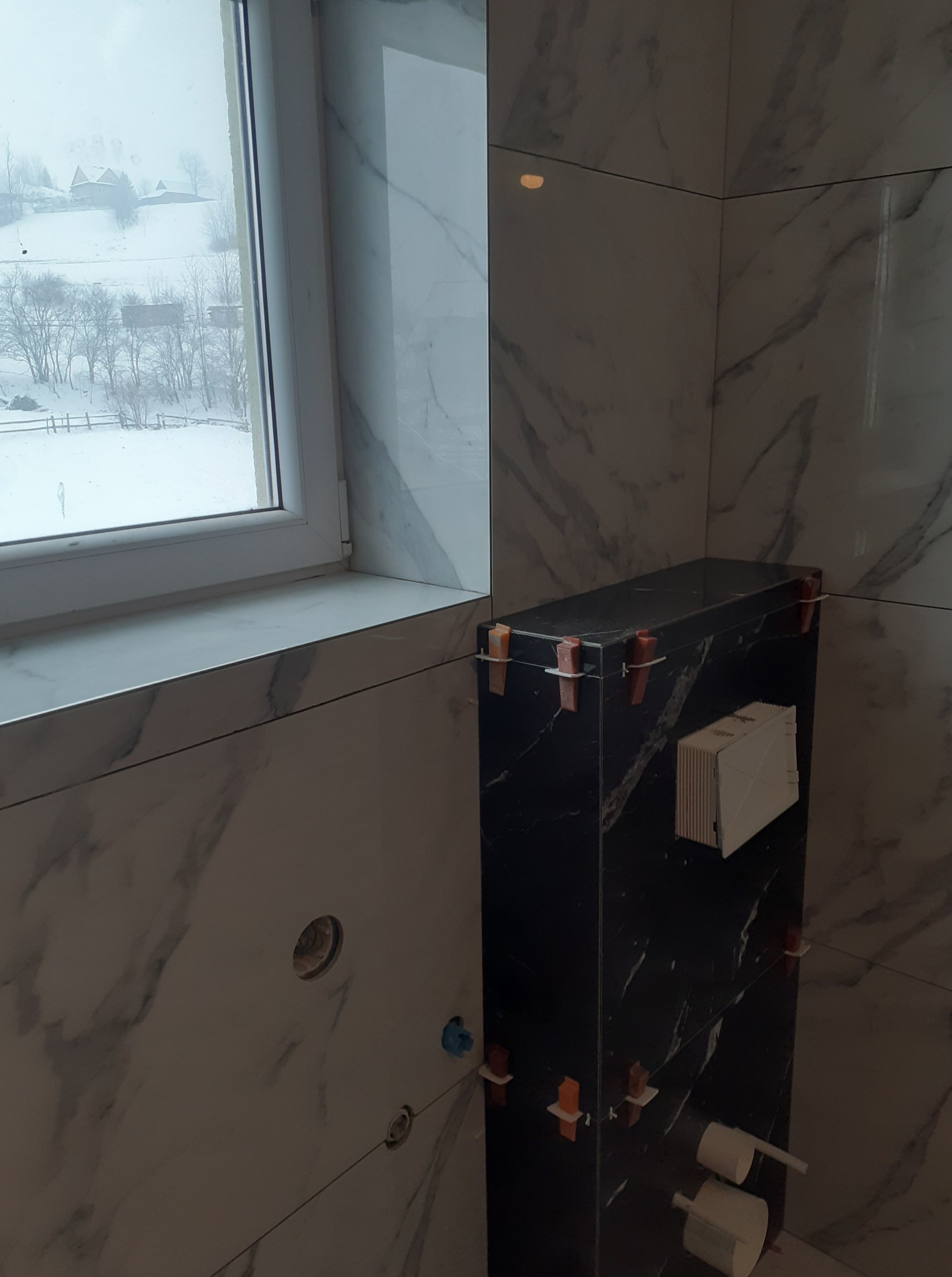 Łazienka w trakcie wykańczania: marmurowe płytki na ścianach, czarny zabudowany stelaż WC z dystansami, widok na zaśnieżony krajobraz przez okno.