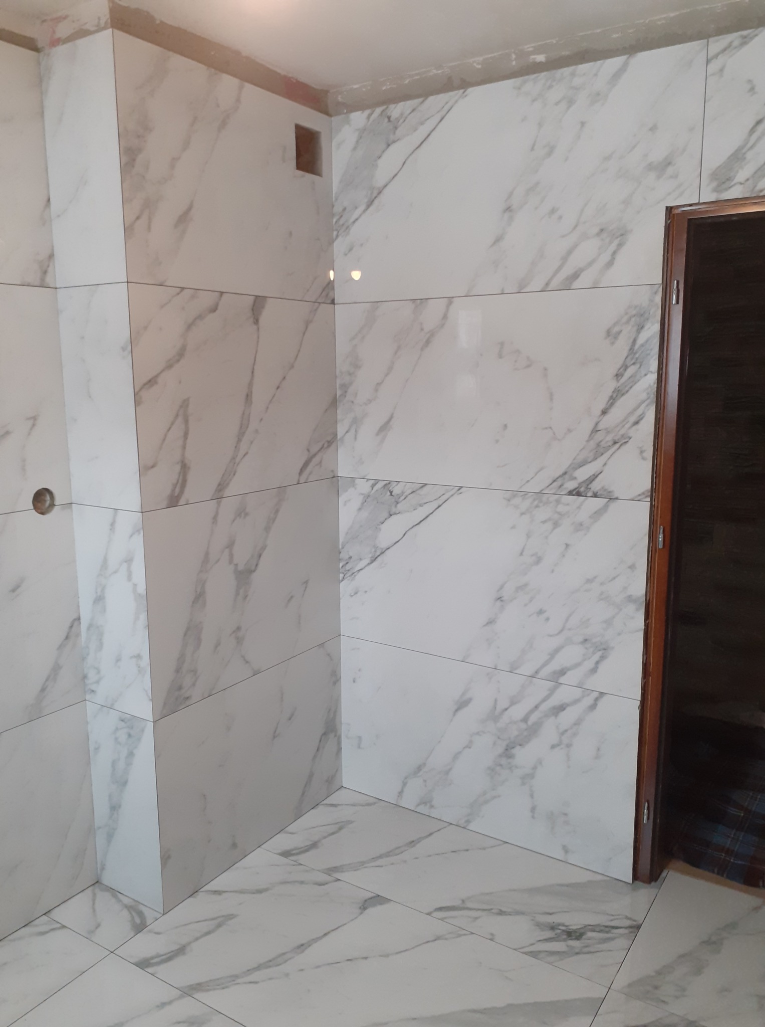 Wykończona łazienka z białymi płytkami imitującymi marmur na ścianach i podłodze. Widoczny fragment futryny drzwi i otwór wentylacyjny w górnej części ściany.