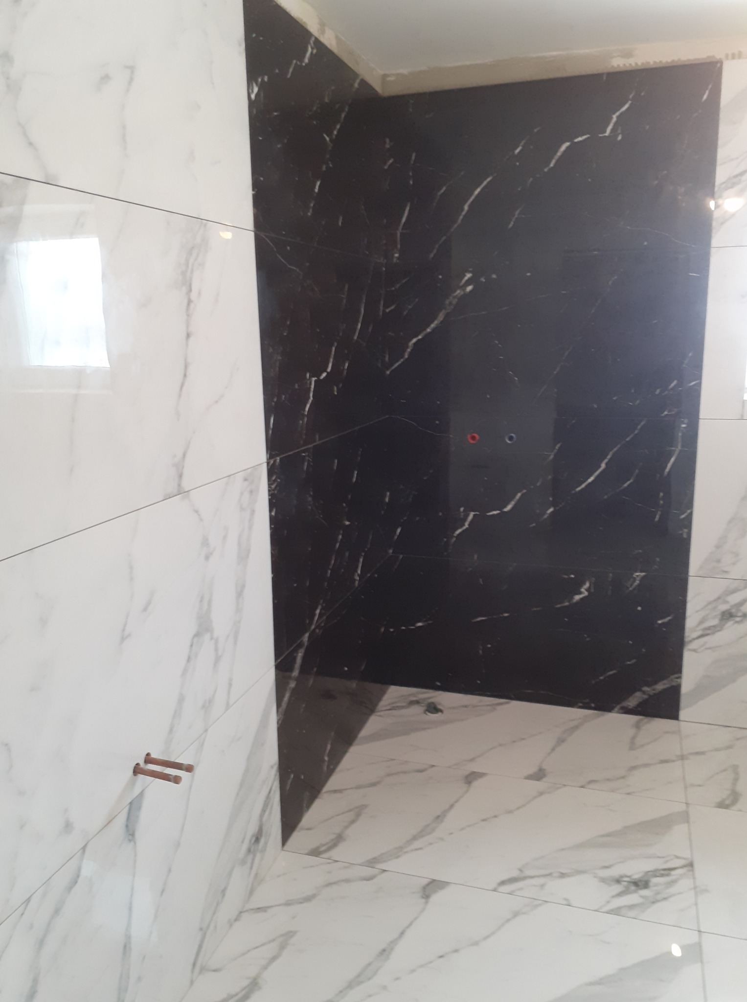 Wykończona łazienka z czarno-białymi płytkami imitującymi marmur na ścianach i podłodze. Widoczne wystające rurki i otwory na baterię.