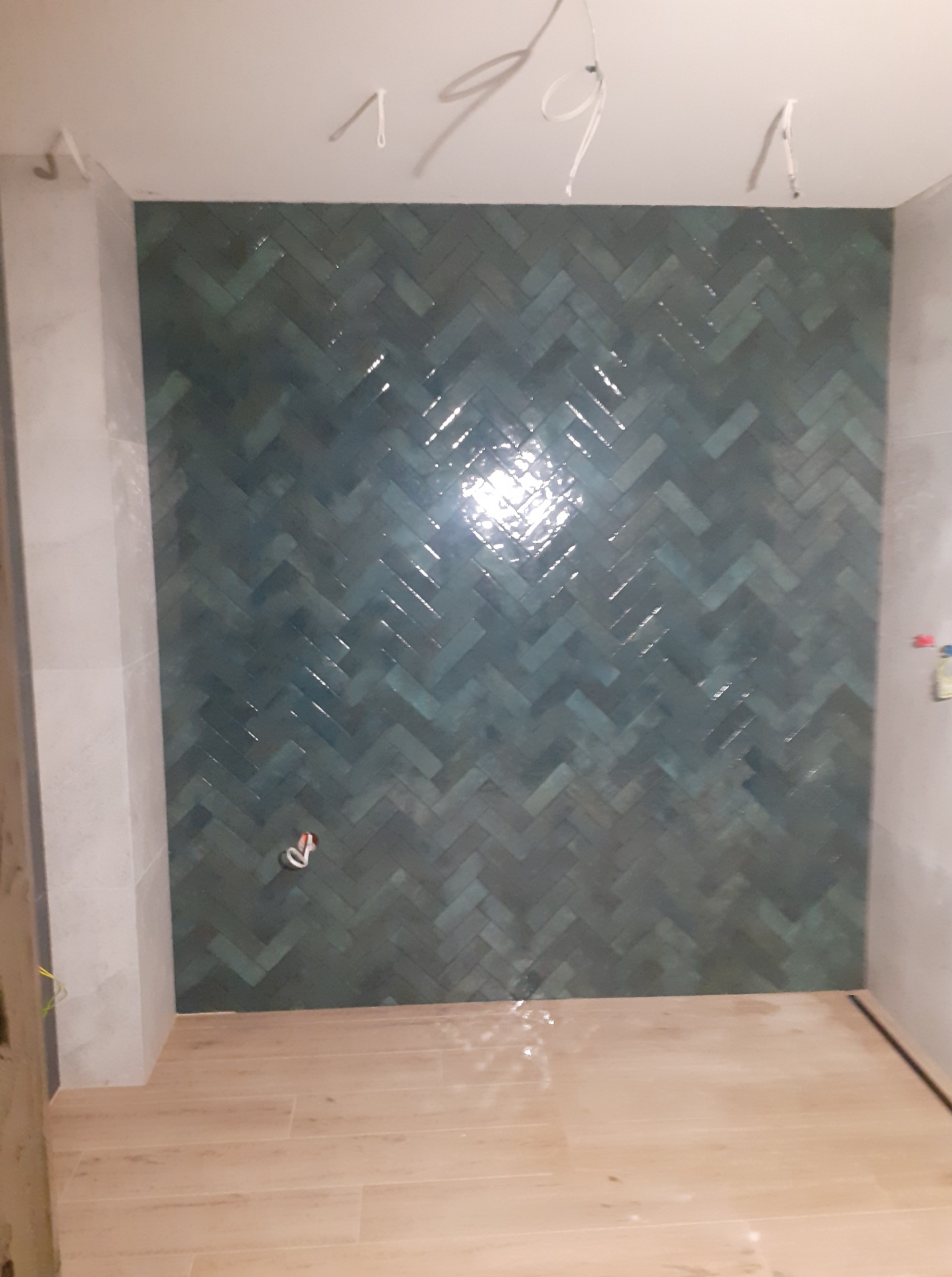 Ściana wyłożona turkusową glazurą w jodełkę, z widoczną instalacją elektryczną na suficie i fragmentem drewnopodobnej podłogi, w trakcie wykańczania łazienki.