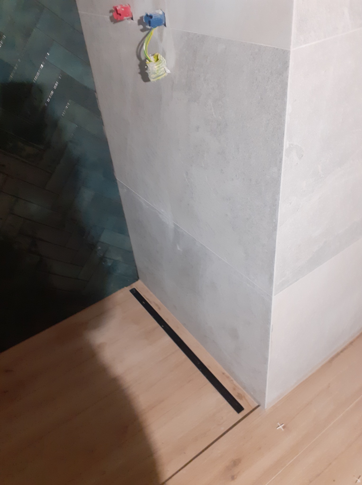 Wykończona ściana z szarych płytek, z widocznymi puszkami elektrycznymi i fragmentem podłogi z jasnego drewna, oddzielonej czarną listwą. W tle ciemne, ułożone w jodełkę płytki.