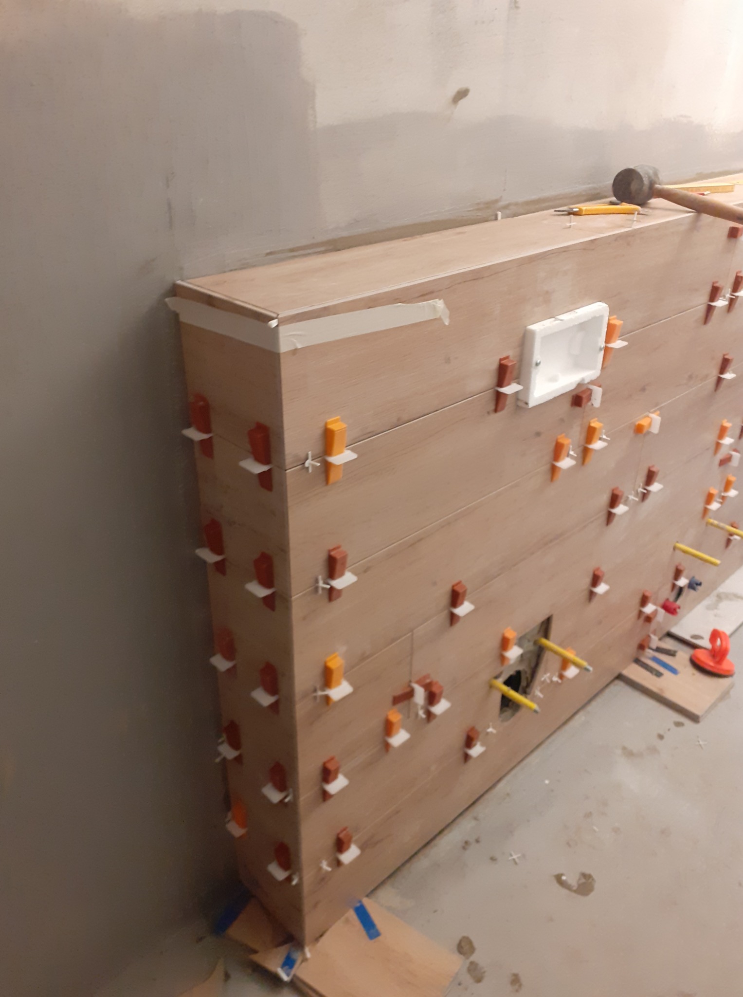 Montaż płytek imitujących drewno na ścianie z użyciem klipsów poziomujących. Widoczny otwór na instalację i narzędzia glazurnicze.