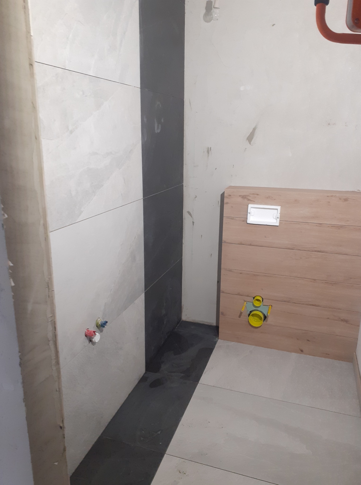 Łazienka w trakcie wykańczania: jasne i grafitowe płytki na ścianach, zabudowa WC z drewnopodobnych paneli, widoczne przyłącza wodne i kanalizacyjne.