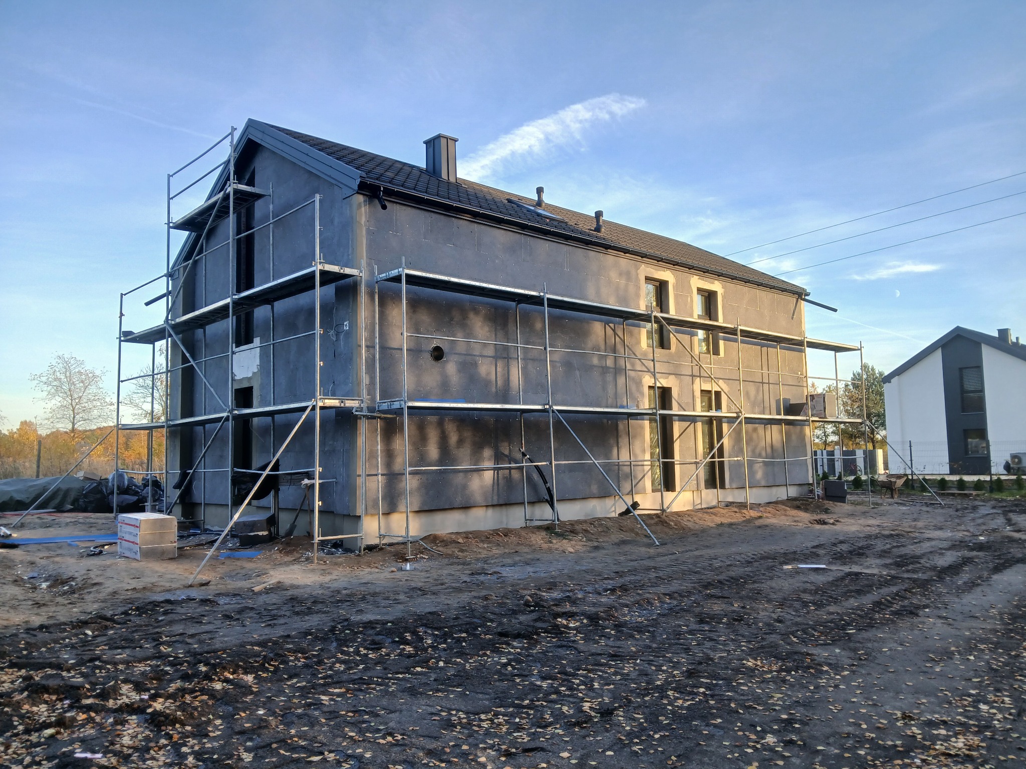 Dom w trakcie prac elewacyjnych, z widocznymi rusztowaniami i ociepleniem ścian. Na pierwszym planie teren budowy z błotem i liśćmi.