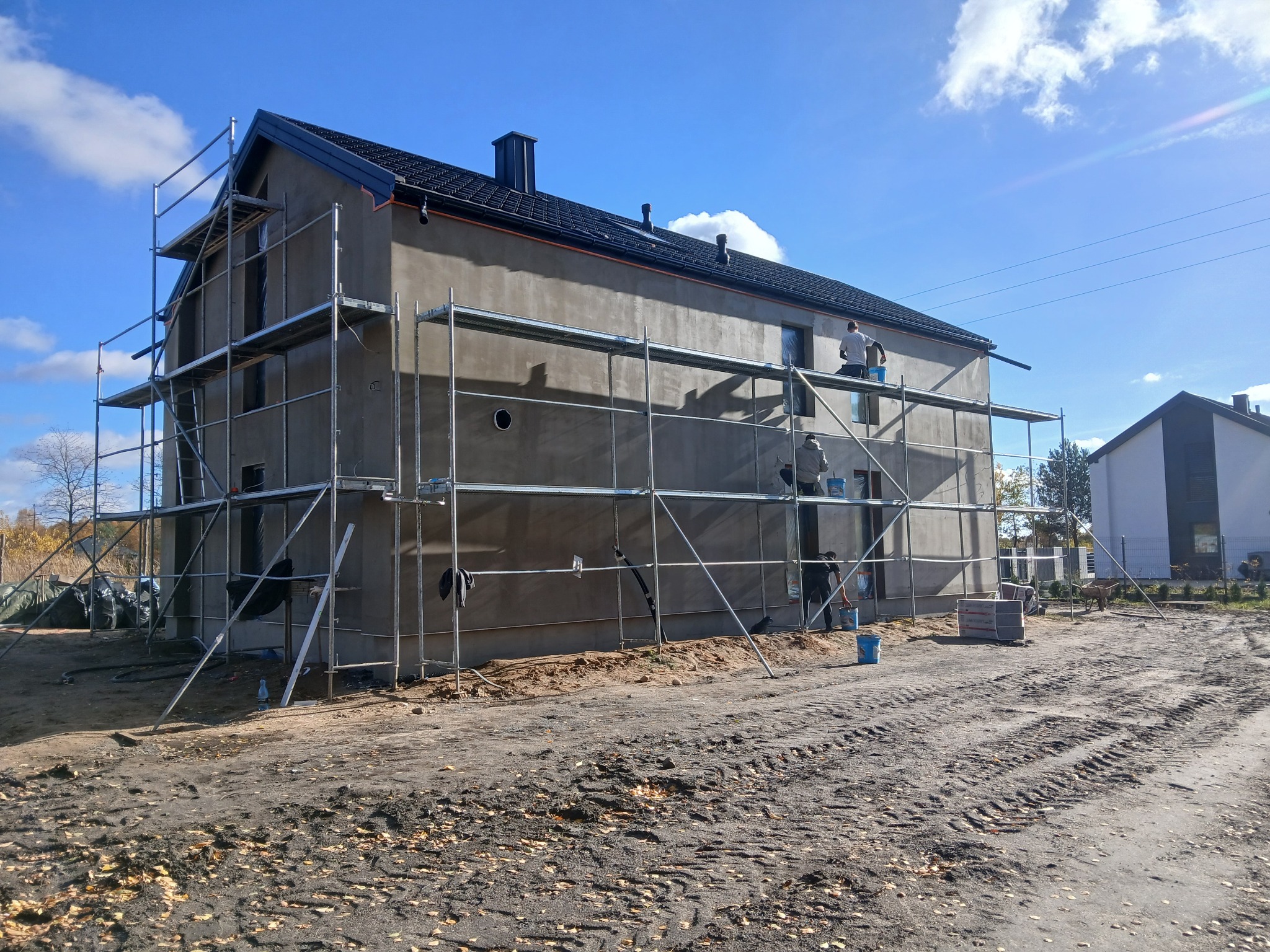 Dwukondygnacyjny dom w trakcie tynkowania elewacji, otoczony rusztowaniem. Widoczni pracownicy w trakcie pracy, błękitne niebo w tle.