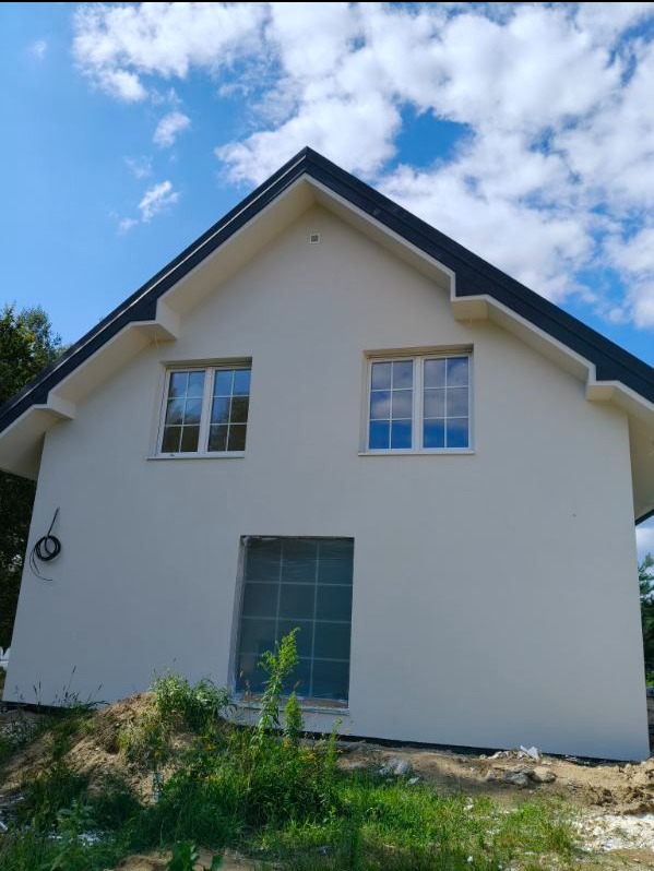 Elewacja domu jednorodzinnego w jasnym kolorze, z oknami i czarnym dachem na tle błękitnego nieba z chmurami. Jedno okno zabezpieczone folią.