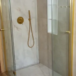 Nowoczesna kabina prysznicowa walk-in z elementami w kolorze złotym, marmurową okładziną i drewnianą podłogą. Wnęka w ścianie.