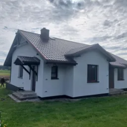 Biały dom z brązowym dachem i oknami, numer '11E' nad oknem. Widoczne schody wejściowe i trawnik. Pochmurne niebo w tle.