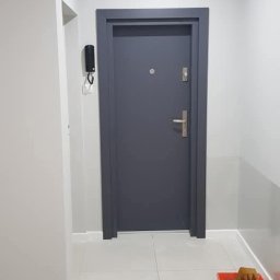 Wymarzony Remont - Szare drzwi wejściowe w odnowionym, pomalowanym na szaro przedpokoju. Na podłodze widoczna kuweta malarska z akcesoriami.