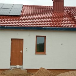 Wymarzony Remont - Dom jednorodzinny z czerwoną dachówką i panelami słonecznymi. Elewacja biała z drewnianymi akcentami. Okna i drzwi w kolorze drewna.