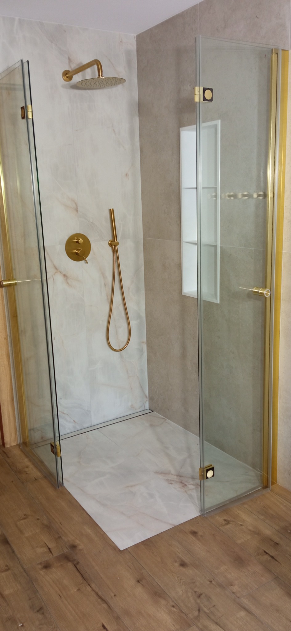 Nowoczesna kabina prysznicowa walk-in z elementami w kolorze złotym, marmurową okładziną i drewnianą podłogą. Wnęka w ścianie.