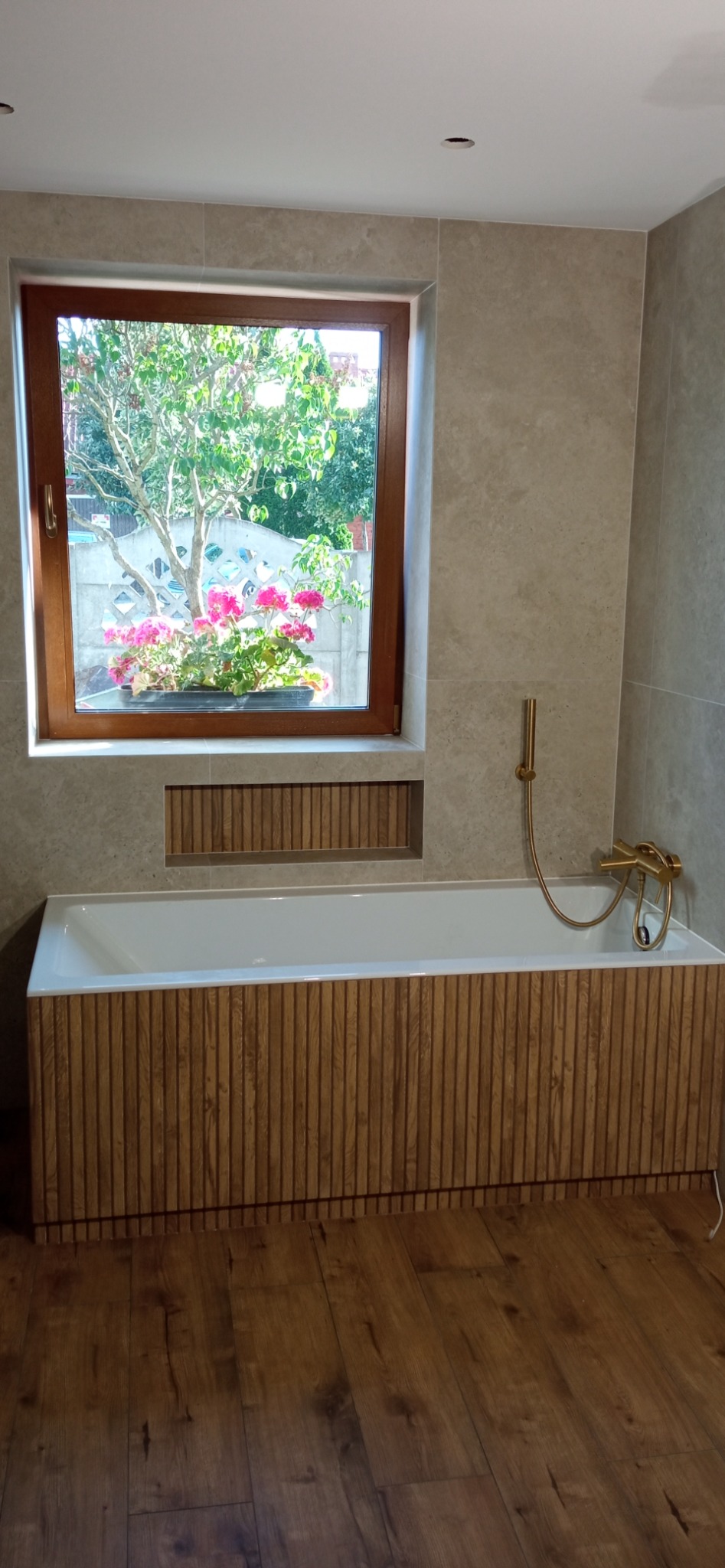 Nowoczesna łazienka z drewnianą obudową wanny, beżowymi płytkami i oknem z widokiem na ogród. Złota armatura dodaje elegancji wnętrzu.