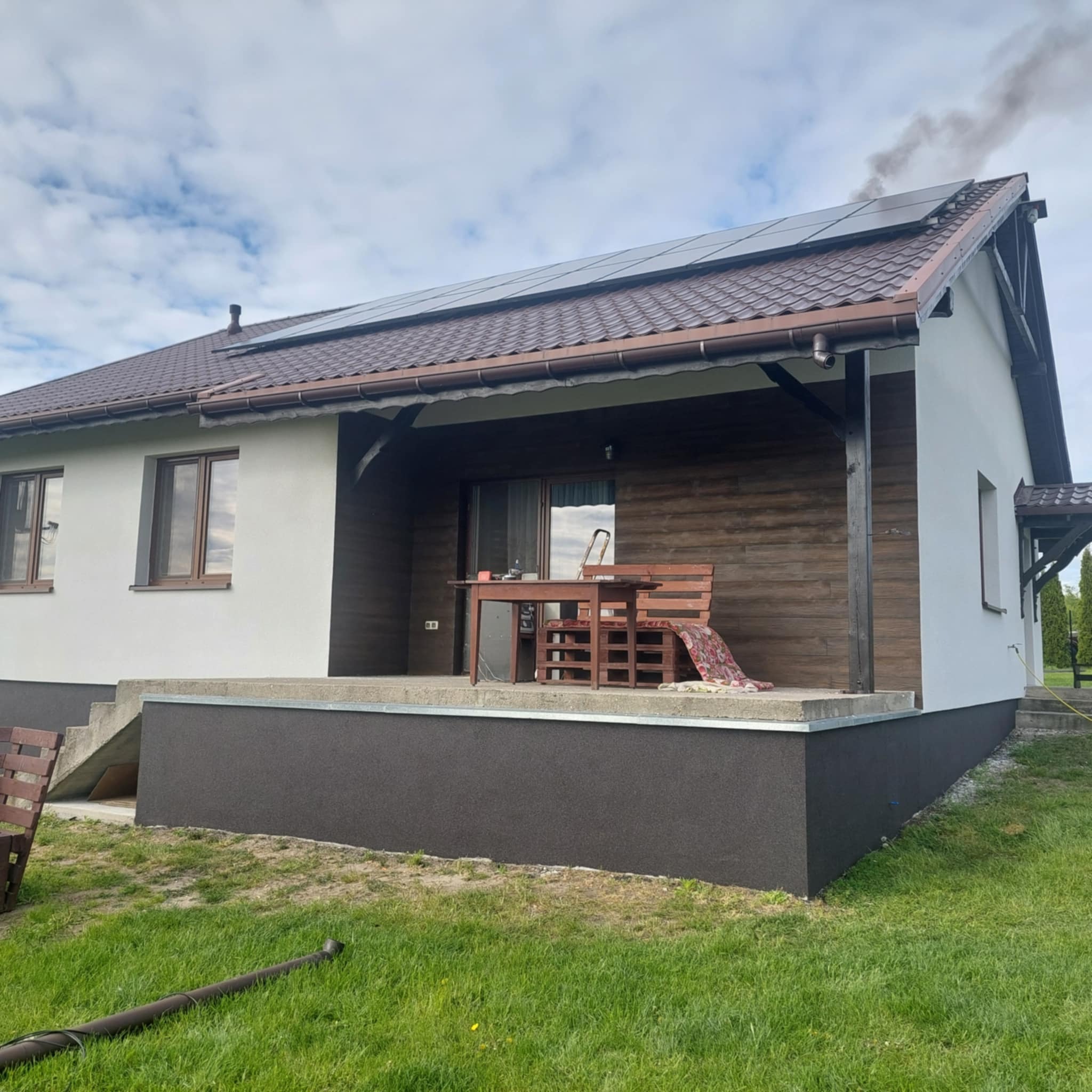 Elewacja domu jednorodzinnego po renowacji: białe ściany, brązowy cokół, drewniana weranda i panele słoneczne na dachu. Widoczne ślady prac wykończeniowych.