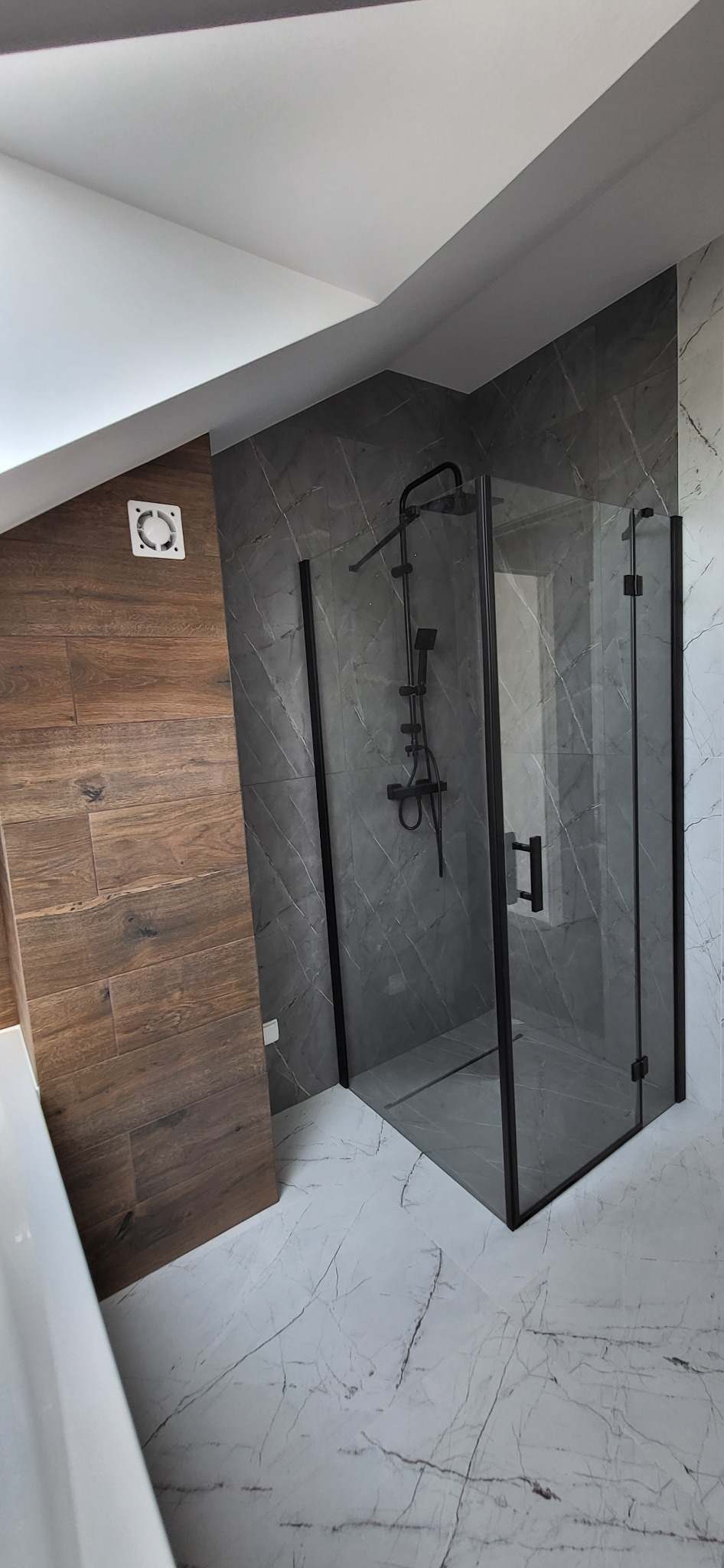 Nowoczesna kabina prysznicowa z czarnymi profilami na tle marmurowych płytek. Widoczny drewnopodobny panel i wentylator sufitowy.