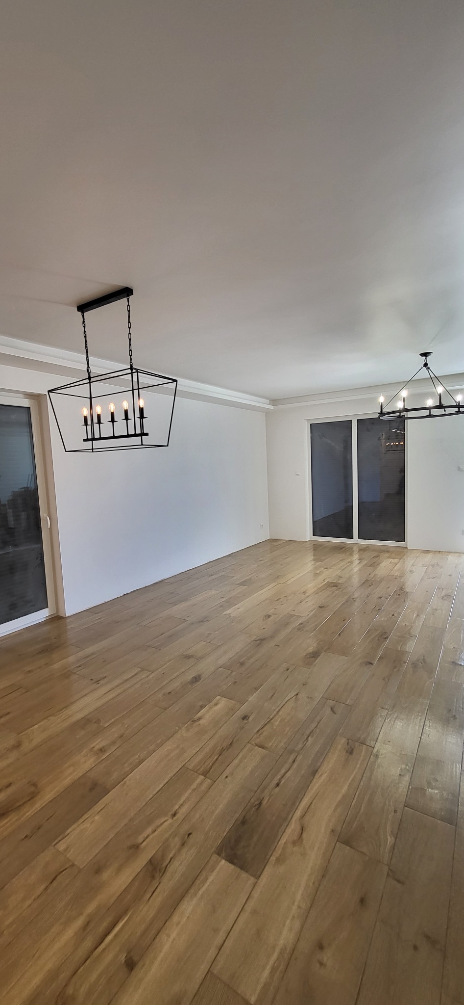 Jasny parkiet w przestronnym salonie z białymi ścianami, listwami i nowoczesnymi lampami. Duże okna przesuwne wpuszczają światło. Widok na świeżo wykończone wnętrze.