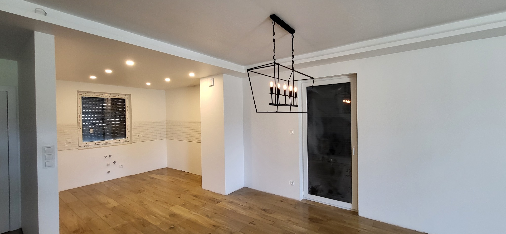 Jasne wnętrze po remoncie: drewniana podłoga, białe ściany z listwami, okno z roletą i nowoczesny, geometryczny żyrandol. Widoczne przygotowania pod montaż wyposażenia.