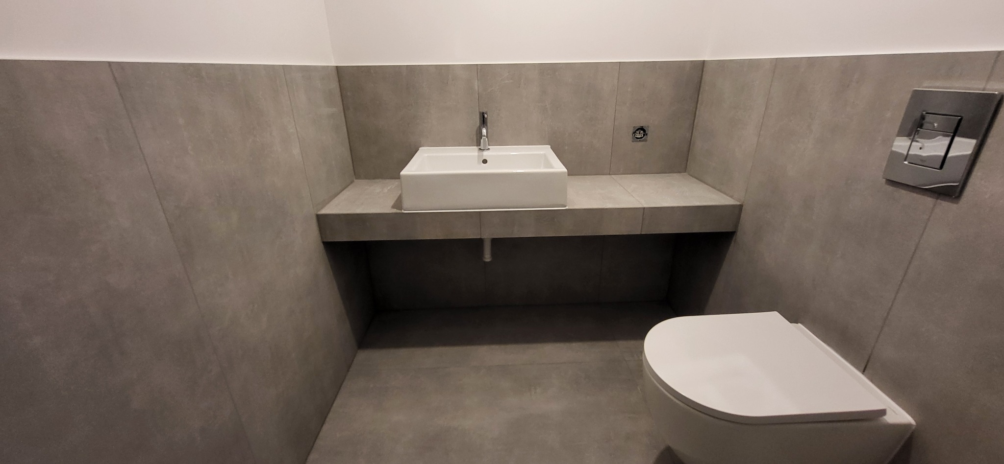 Nowoczesna łazienka z szarymi płytkami, białą umywalką na blacie i podwieszaną toaletą. Minimalistyczny design,  oświetlenie górne,  srebrny przycisk spłuczki.