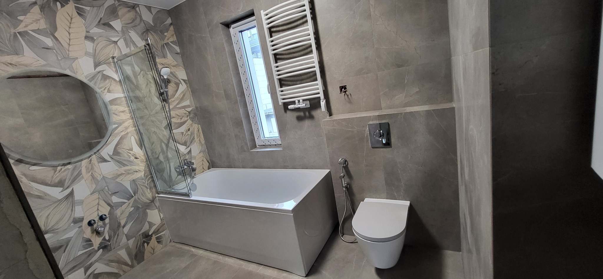 Wykończona łazienka z wanną z parawanem, grzejnikiem drabinkowym i toaletą w szaro-białej kolorystyce. Ściany wyłożone płytkami imitującymi marmur.