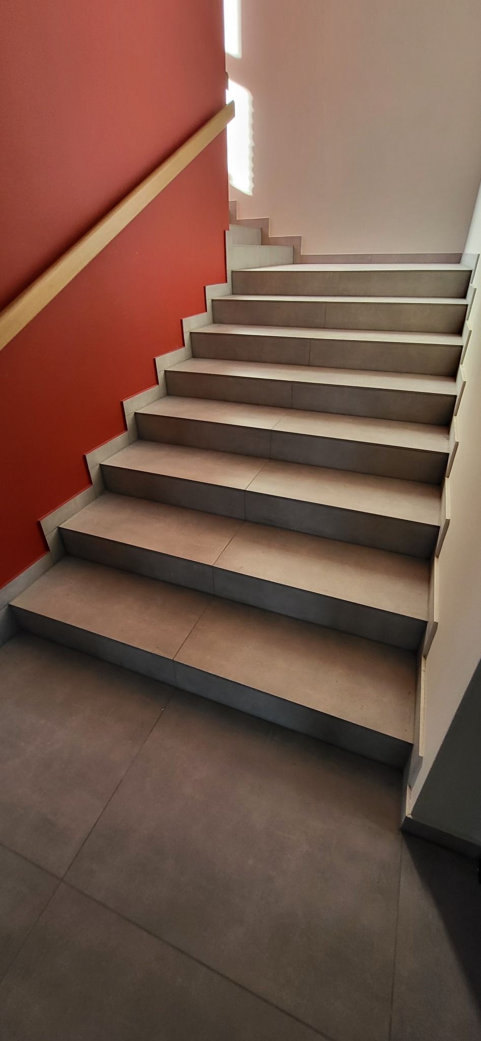 Wykończone schody z płytkami w Przeźmierowie, z drewnianą poręczą na tle czerwonej ściany. Widoczne płytki na stopniach i podłodze, jasne oświetlenie.