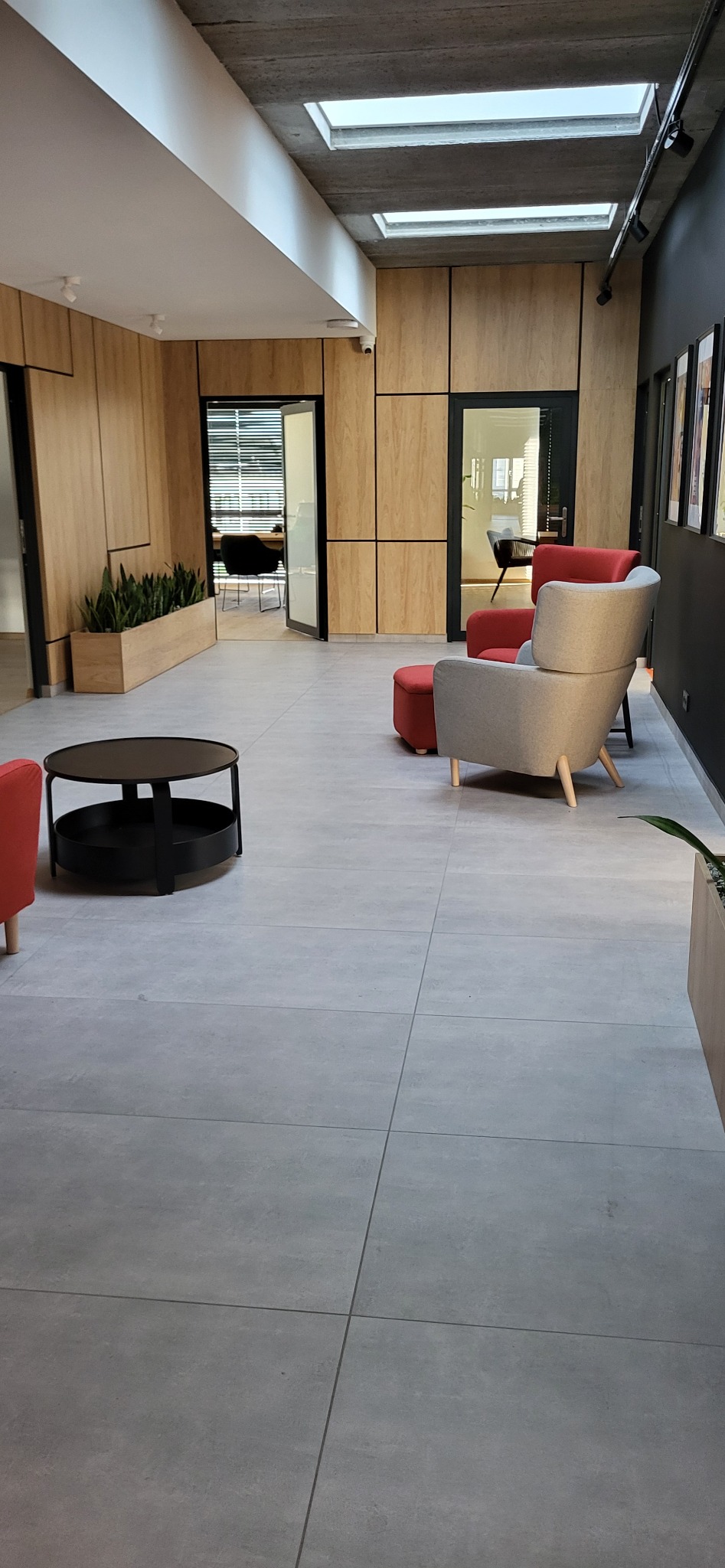 Nowoczesna przestrzeń biurowa z szarymi płytkami podłogowymi, drewnianymi panelami ściennymi i designerskimi fotelami w odcieniach szarości i czerwieni, doświetlona naturalnym światłem.