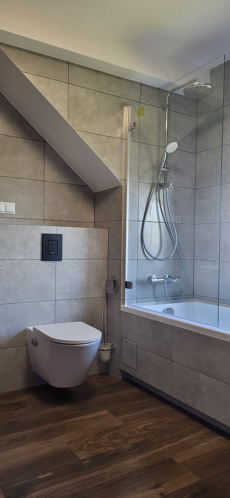 Nowoczesna łazienka w szarościach: płytki ścienne, drewnopodobna podłoga, biała toaleta podtynkowa z czarnym przyciskiem i kabina prysznicowa ze szklaną ścianką.